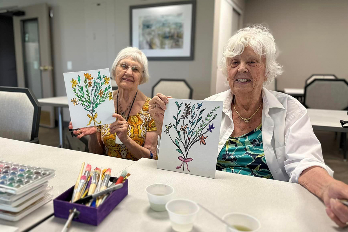 Two memory care residents are holding up paintings of foliage that they created together. 