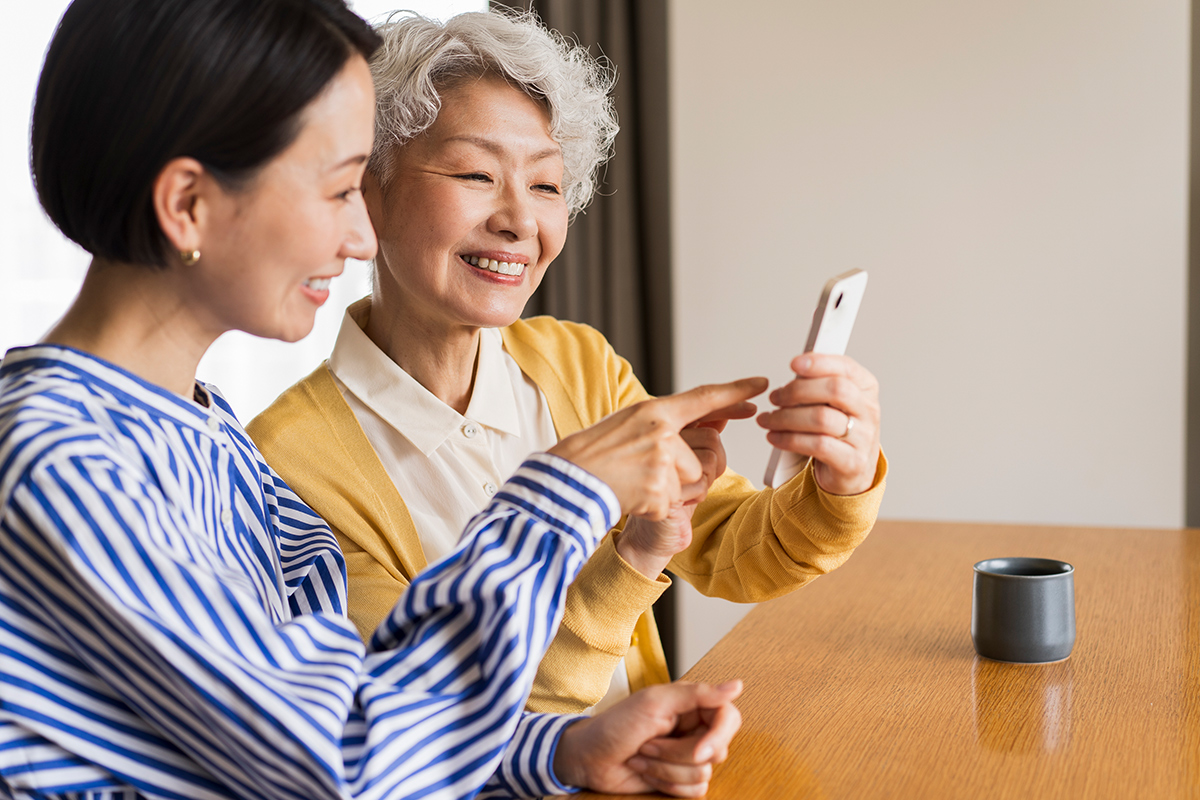 A senior woman and her adult child both looking at a smartphone and smiling. 