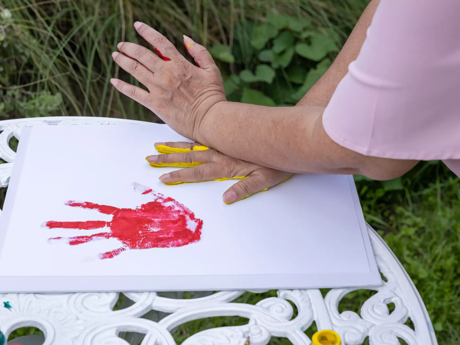 A senior woman's hands, previously dipped into red paint, are pressing into a pad of paper and leaving behind a red handprint.