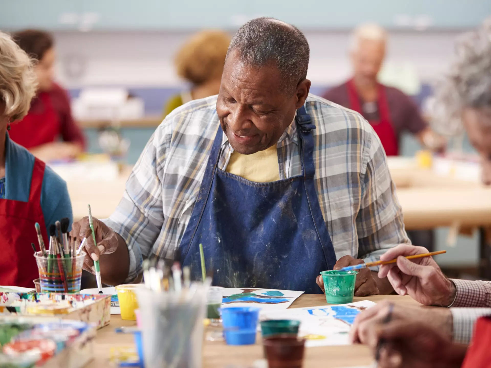 A senior man wearing an art apron, seated at a table amongst other seniors. He is focused on painting on a piece of paper in front of him.