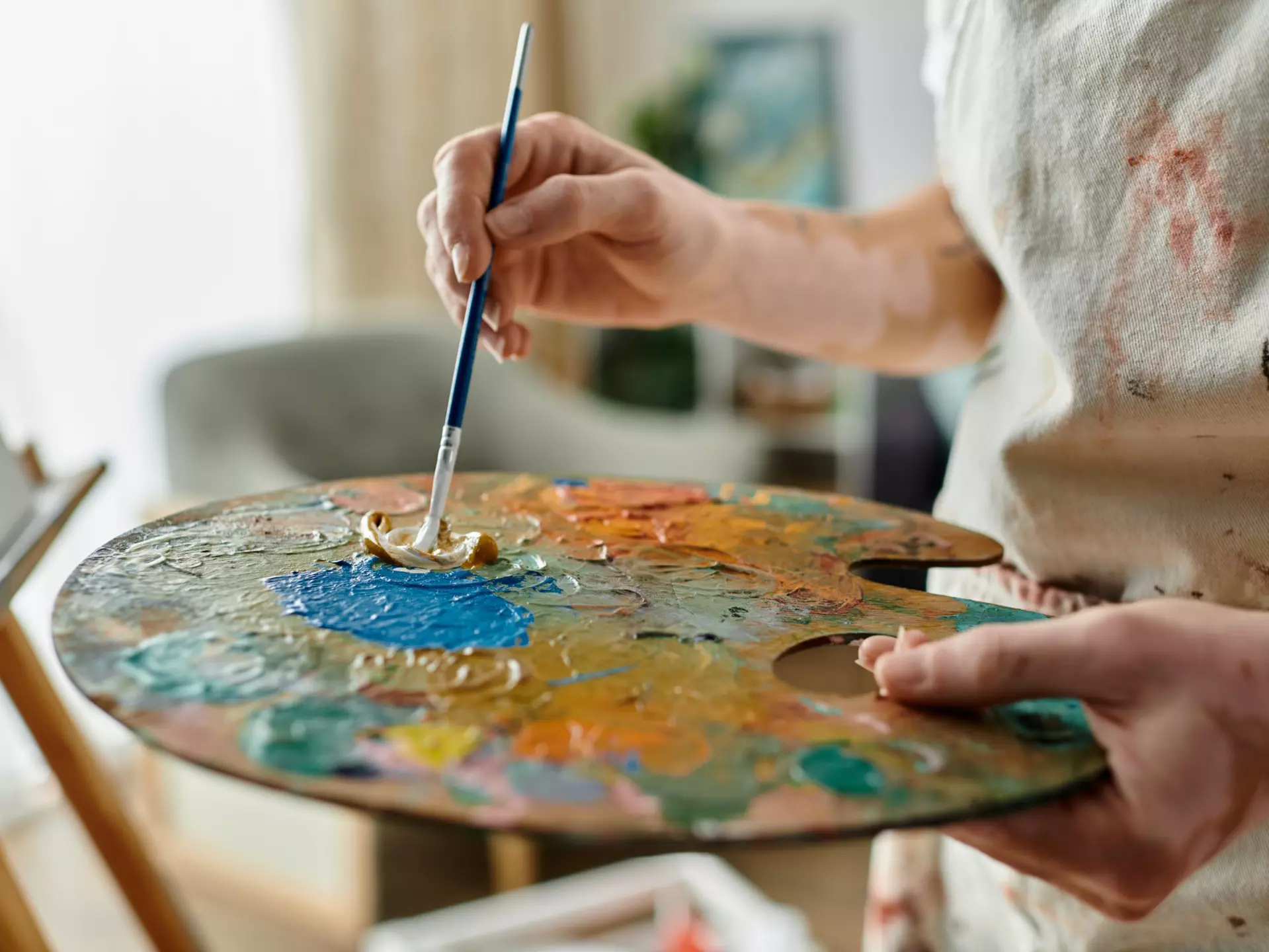 An artist's paint palette with different shades of paint applied. A hand holding a paintbrush is dipping into one of the shades of paint.
