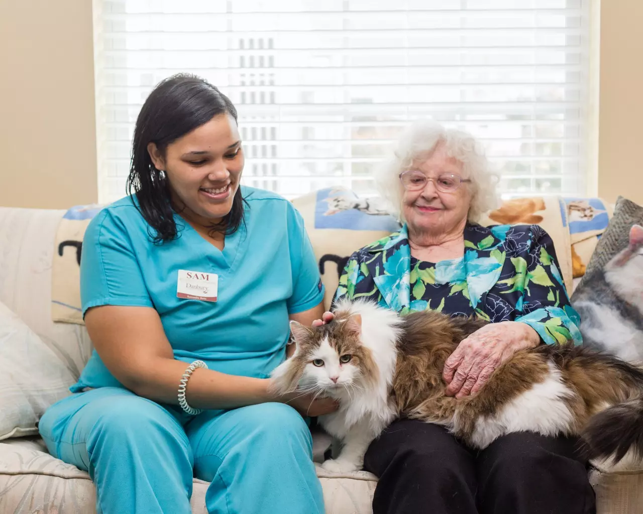 A caregiver and a StoryPoint Group resident are sitting on a sofa, with a cat on the residents lap. 