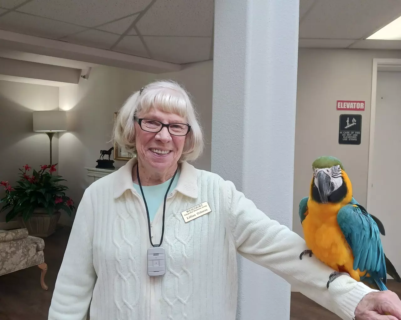 A happy StoryPoint Group resident is standing with her arm outstretched, with a colorful parrot perched on her arm.