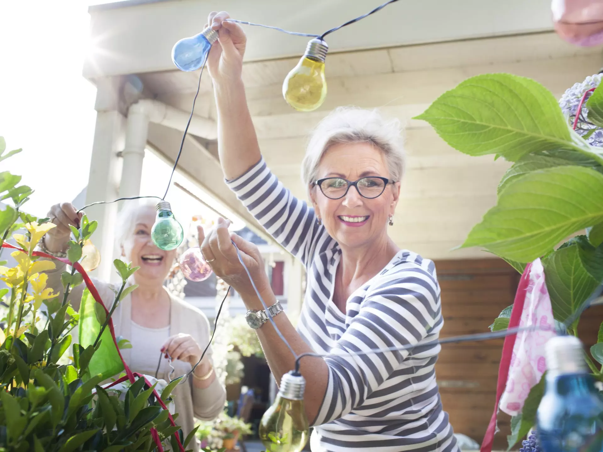 Two older women in garden, hanging colorful lights.