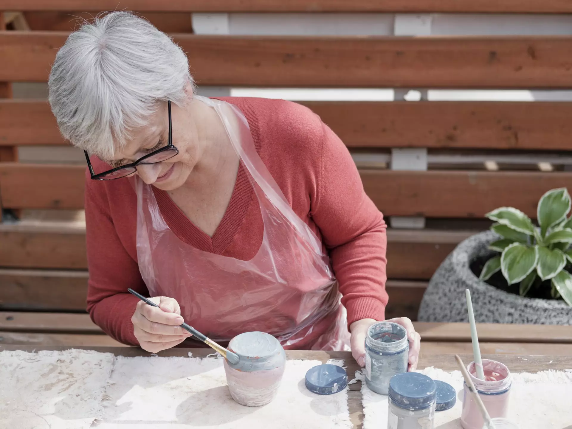 Older gray haired woman with glasses painting handmade clay vase with colored glaze using a brush.