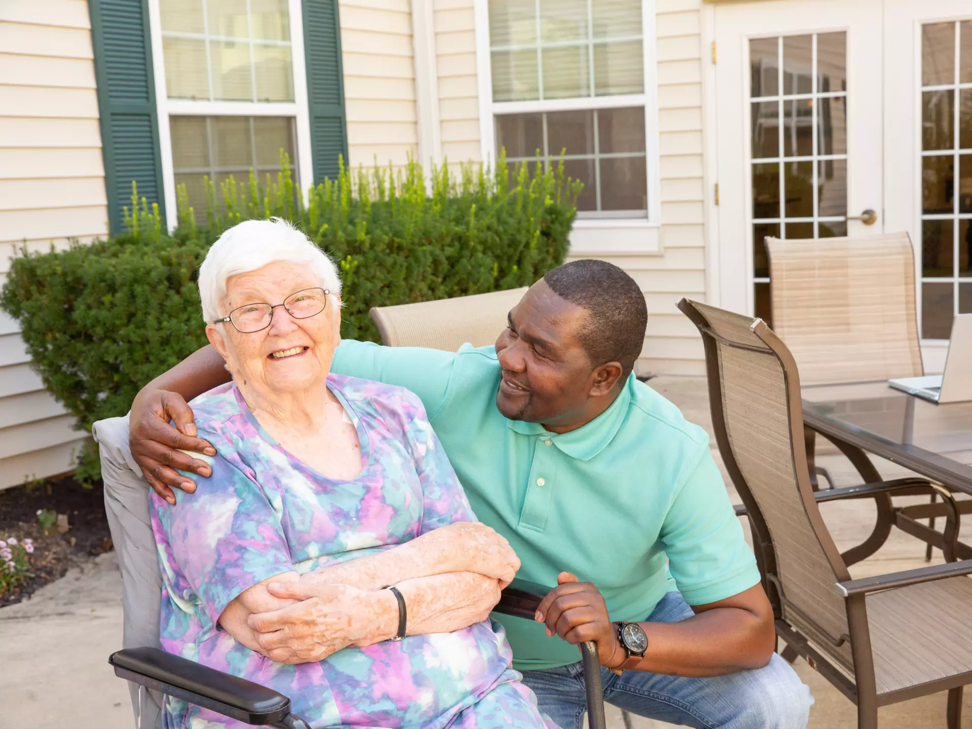 A caregiver and senior are happily lounging at an outdoor patio area. 