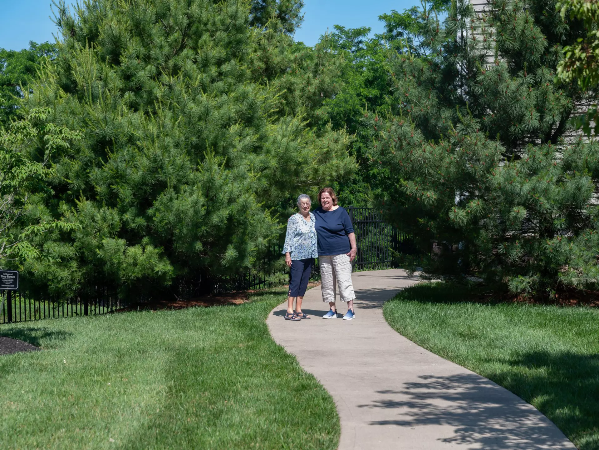 Two senior women pose for a photo at the end of a scenic outdoor walking path surrounded by greenery.